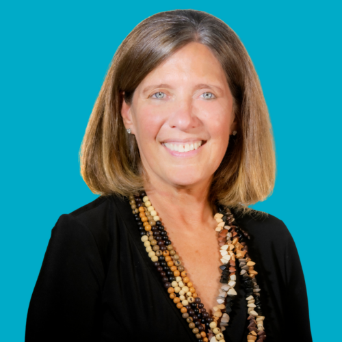 A woman with shoulder-length brown hair, wearing a black top and a multicolored beaded necklace, smiles in front of a solid turquoise background.
