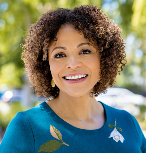 Woman with curly hair wearing a teal top with floral embroidery, smiling outdoors with greenery in the background.