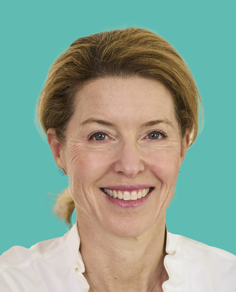 Smiling woman with light brown hair pulled back, wearing a white shirt, in front of a solid teal background.