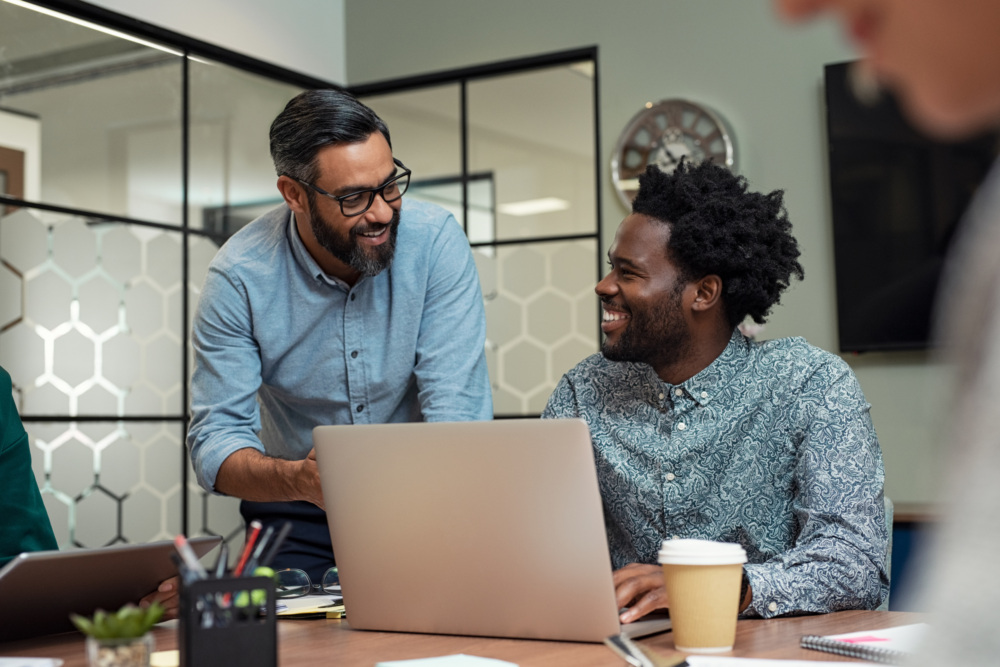 Two men in an office setting smile and talk while working at a desk with laptops, notebooks, and a coffee cup.