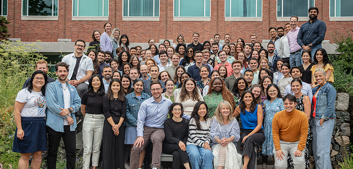 A large group of people poses together outdoors in front of a brick building, smiling at the camera.