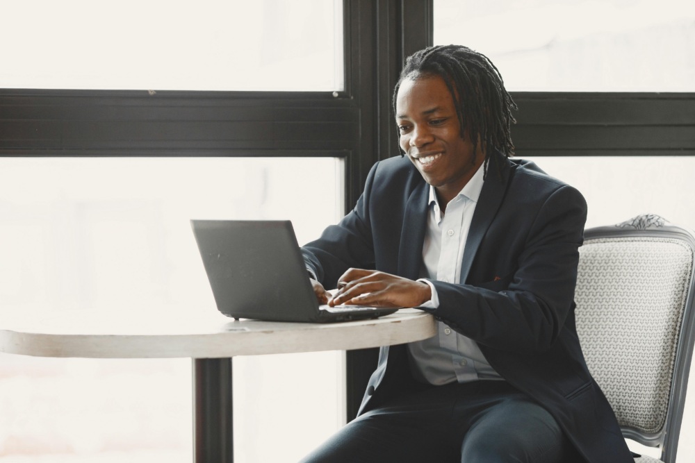 A person in a suit sits at a round table, typing on a laptop and smiling, with large windows in the background.