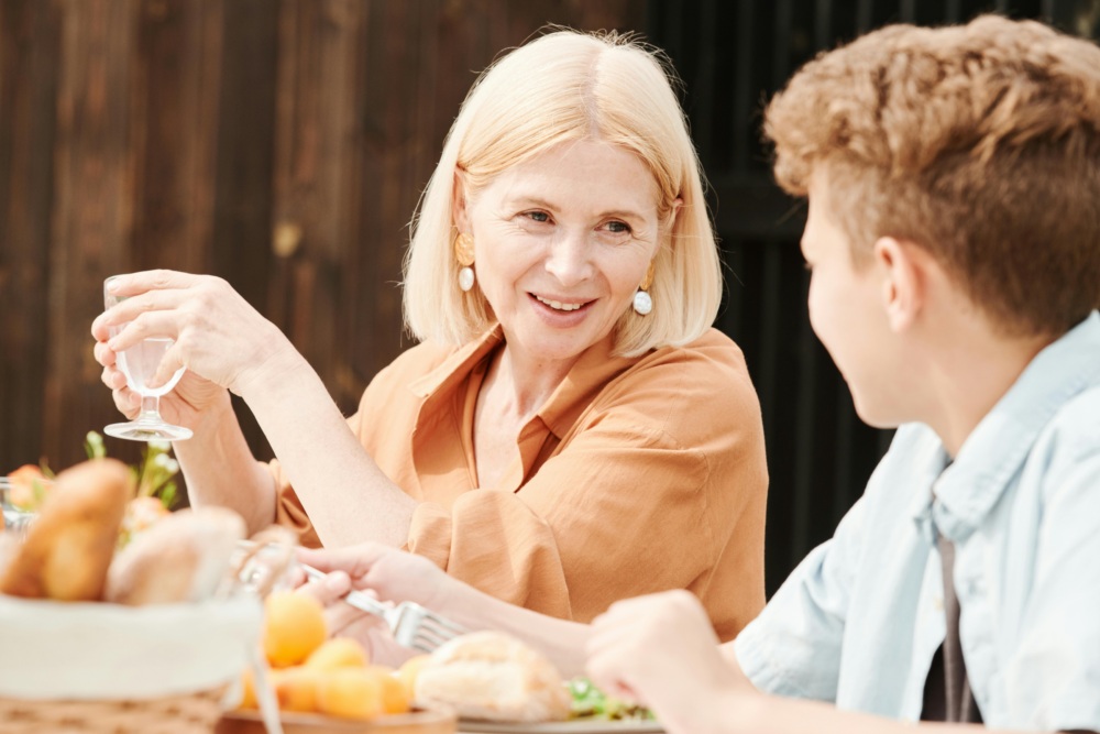 Older woman and young boy sit at an outdoor table with food, smiling and talking to each other during a meal.