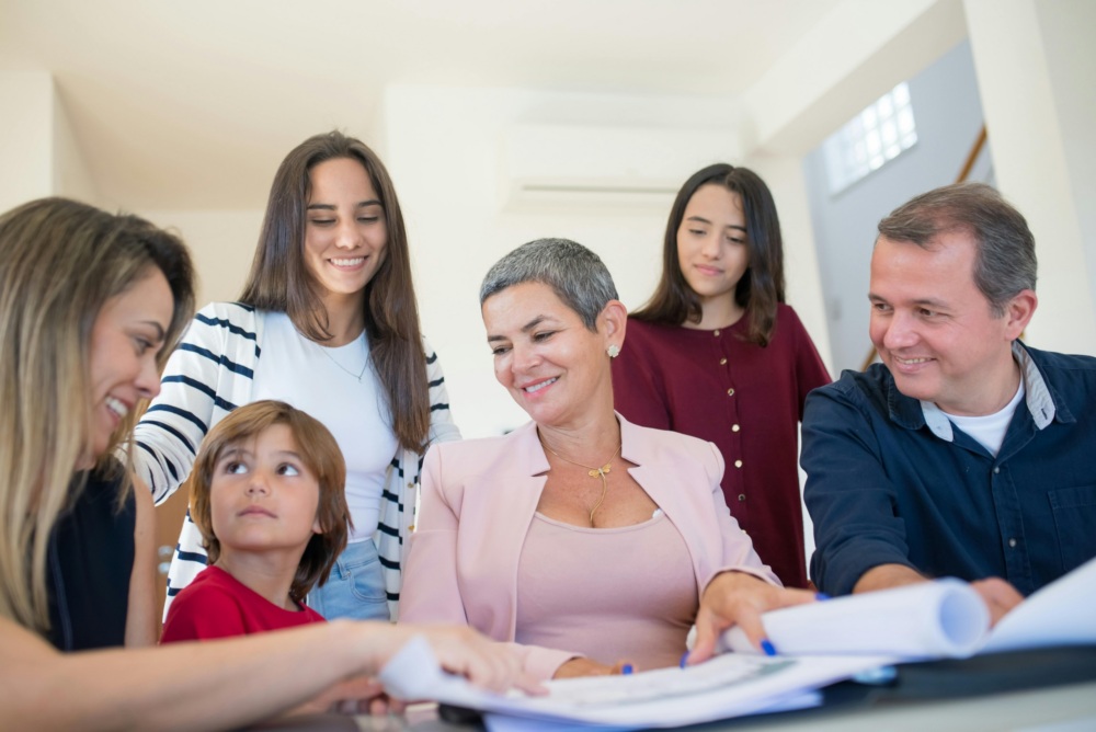 Six people, including adults and children, sit and stand around a table with documents, appearing to discuss or review paperwork together in a well-lit room.