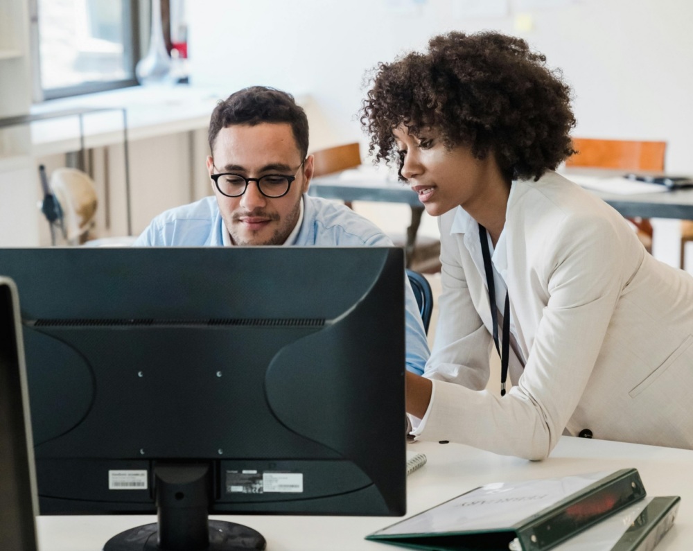 Two colleagues look at a computer monitor together in an office. One person sits at the desk while the other stands next to them, both focused on the screen.