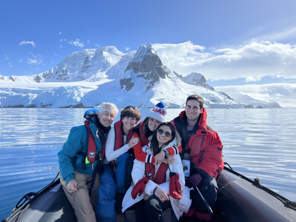 Five people in winter clothing sit together on a small boat with snow-covered mountains and calm water in the background.