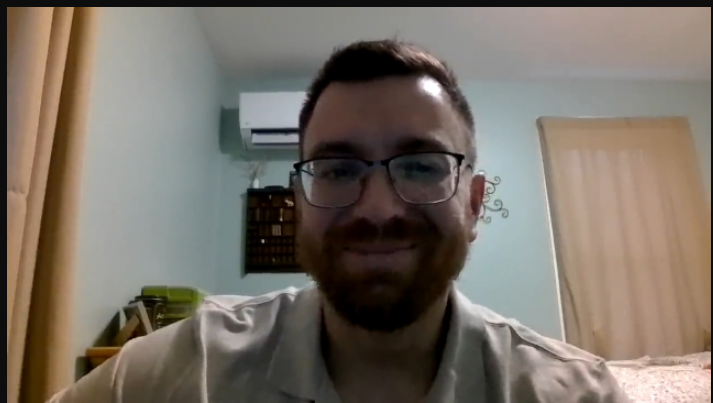 A man with glasses and a beard smiles at the camera in a room with light-colored walls, a window with beige curtains, and an air conditioning unit on the wall.