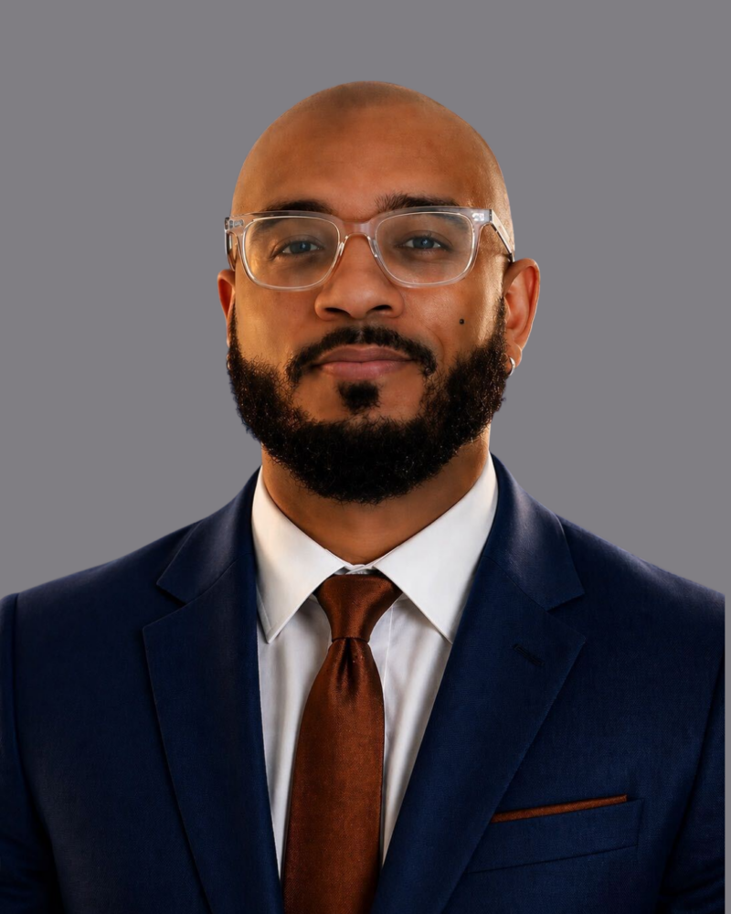 A man with a beard and glasses wearing a navy suit, white shirt, and brown tie, posing against a plain gray background.