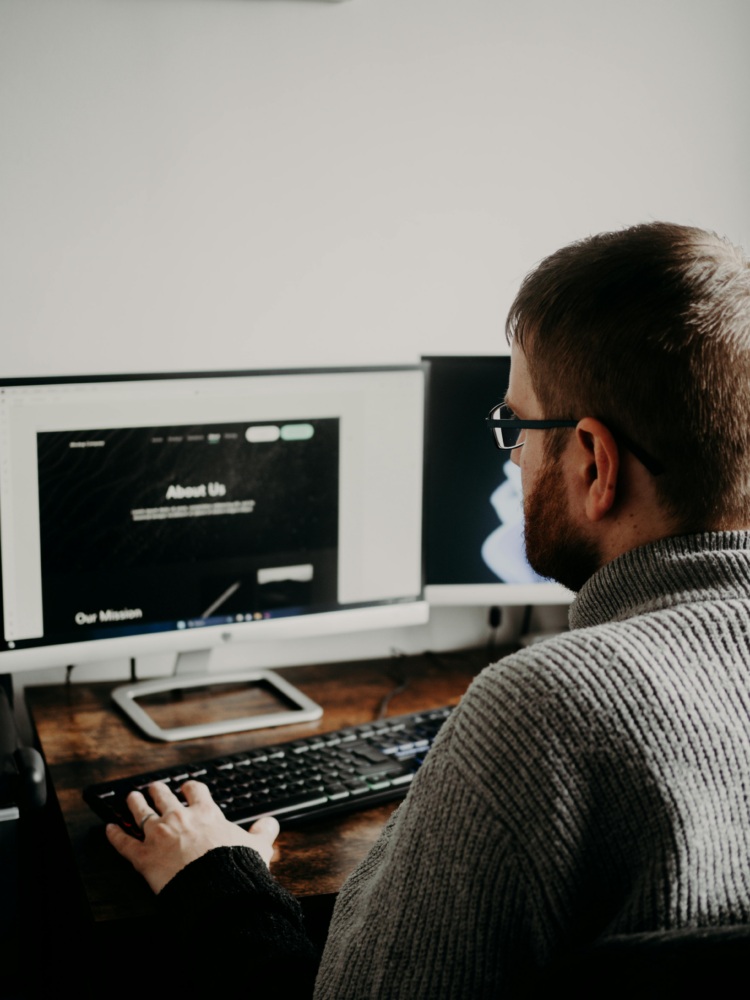 A person wearing glasses and a gray sweater uses a desktop computer at a wooden desk, viewing a website with an "About Us" section on the monitor.