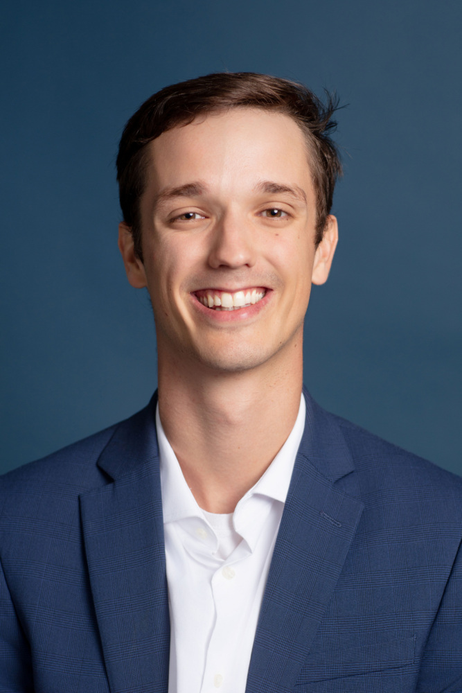A young man with short brown hair smiles, wearing a navy blue suit jacket and white shirt, posed against a plain blue background.