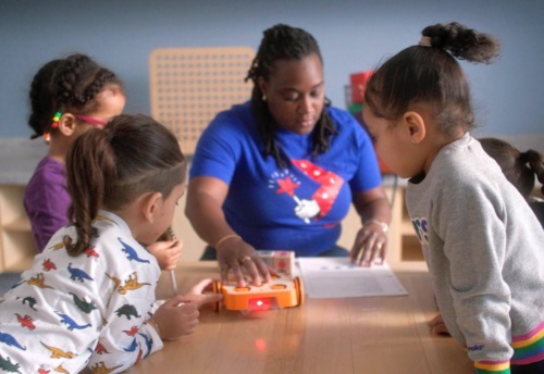 An adult shows four children how to use a small orange robot on a table in a classroom setting.