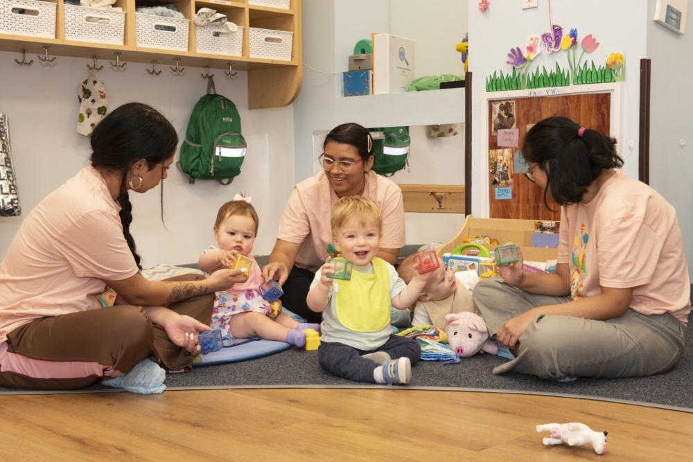 Two adults and two toddlers play with toys on the floor of a daycare classroom. The room has cubbies, backpacks, and colorful decorations on the walls.
