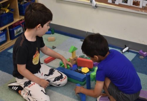 Two young children sit on a blue carpet and build a colorful structure with foam blocks in a classroom.