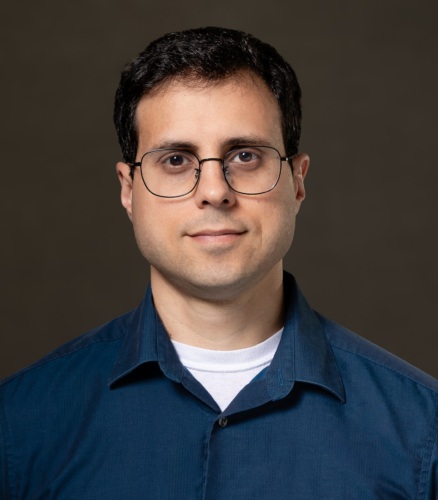 Man with short dark hair and glasses wearing a blue button-up shirt over a white t-shirt, facing the camera with a neutral expression against a plain dark background.