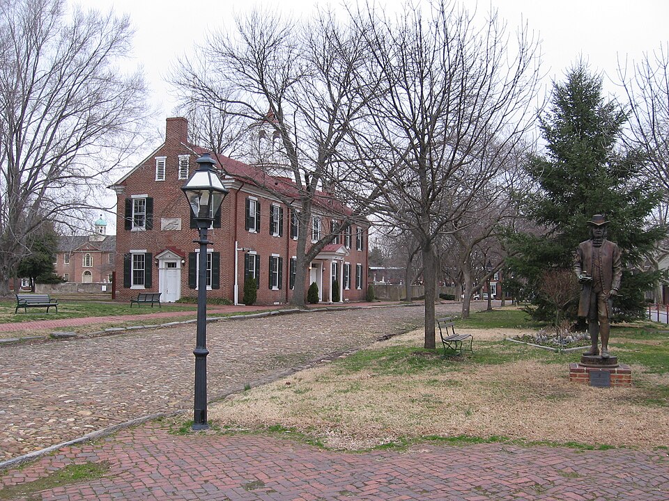 A brick building stands by a cobblestone street lined with leafless trees, a vintage lamppost, and a statue of a man in a suit and hat on a cloudy day.