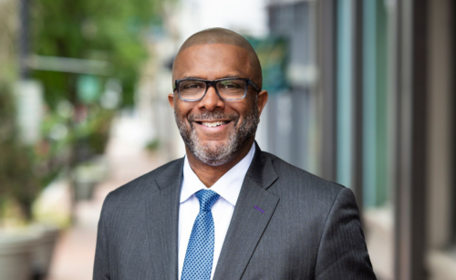 A man wearing glasses, a dark suit, and a blue tie stands on a city sidewalk, smiling with one hand in his pocket.