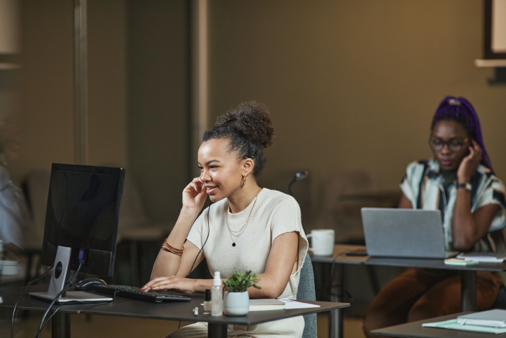 Two women work at desks; one speaks on a headset while using a desktop computer, and the other types on a laptop.
