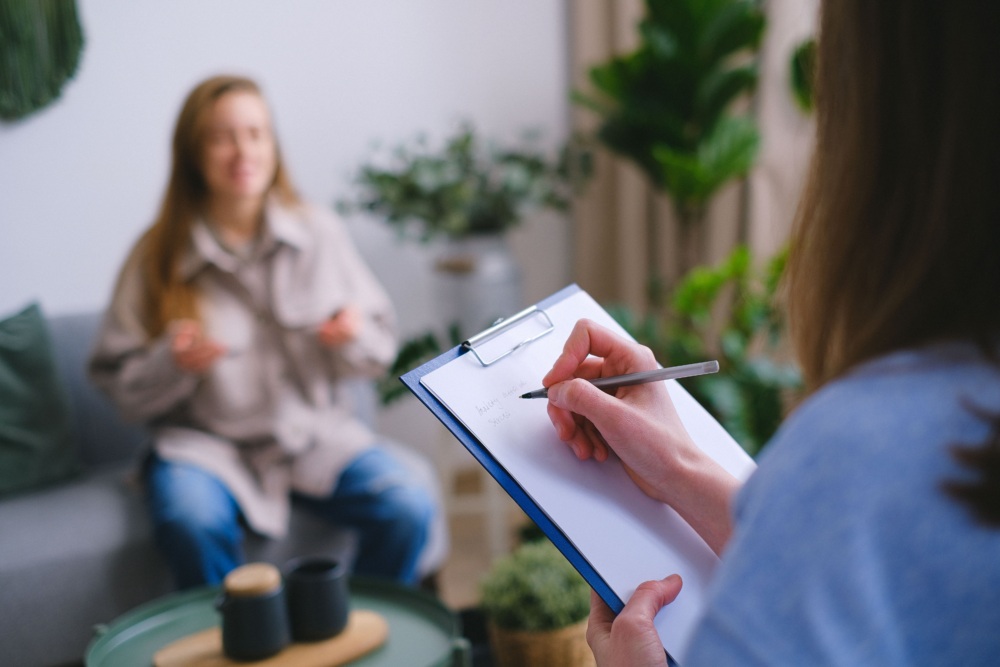 A person writes on a clipboard while another person sits on a couch in the background, speaking with hand gestures.
