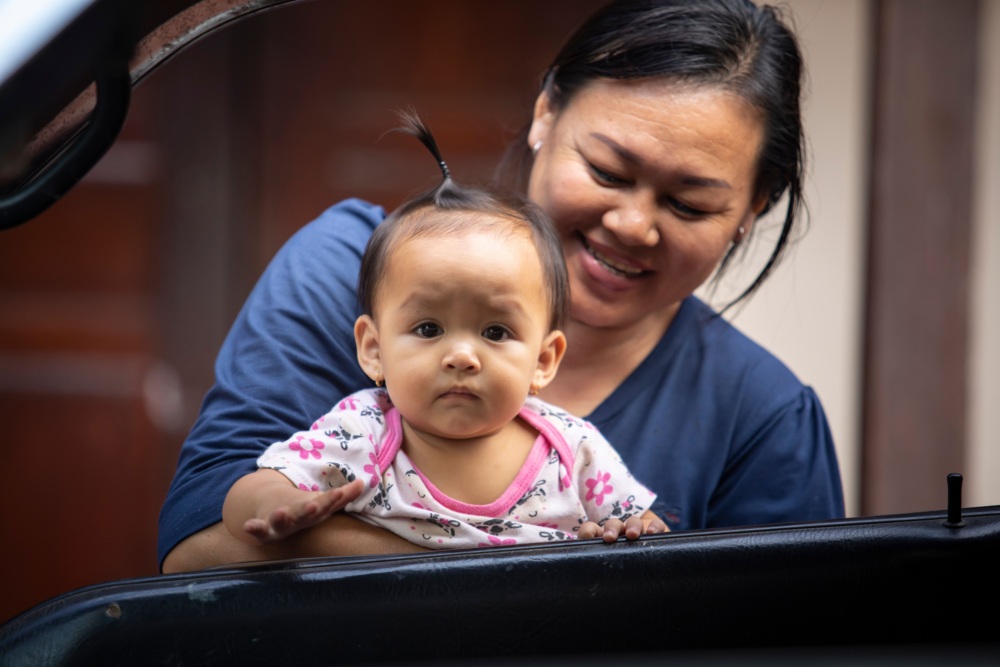A woman smiles while holding a baby with a small ponytail, who looks forward with a neutral expression. They are near a car door.