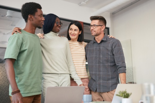 Four people stand together in an office, smiling and looking at each other, with arms around each other’s shoulders. Laptops and office supplies are visible on the desk in front of them.