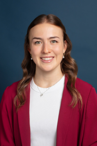 Young woman with long brown hair wearing a white top and red blazer, smiling in front of a plain dark blue background.