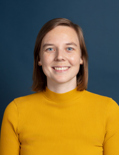 Woman with straight light brown hair wearing a ribbed yellow top, smiling at the camera, photographed against a plain dark blue background.