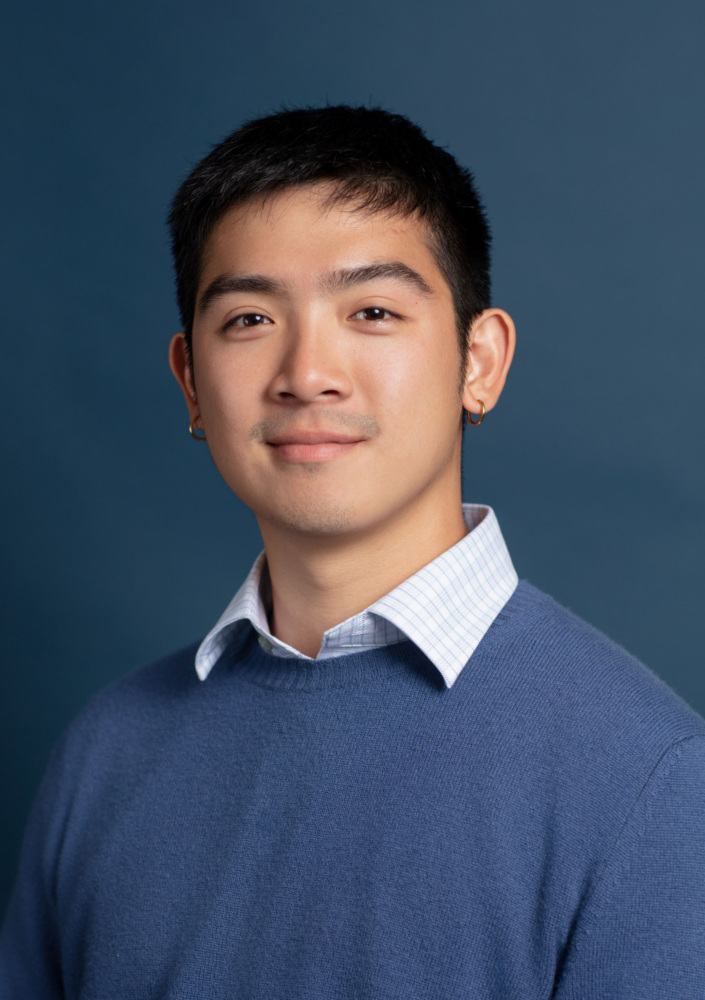 A young person with short dark hair wearing a blue sweater over a collared shirt poses in front of a solid blue background.