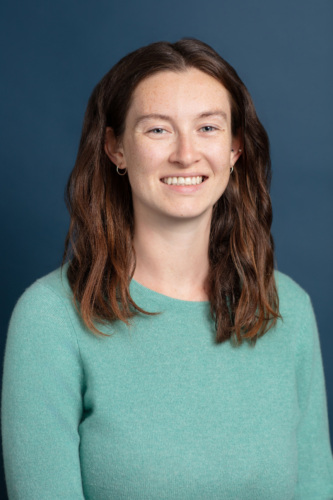 A woman with long brown hair wearing a light green sweater smiles in front of a plain dark blue background.