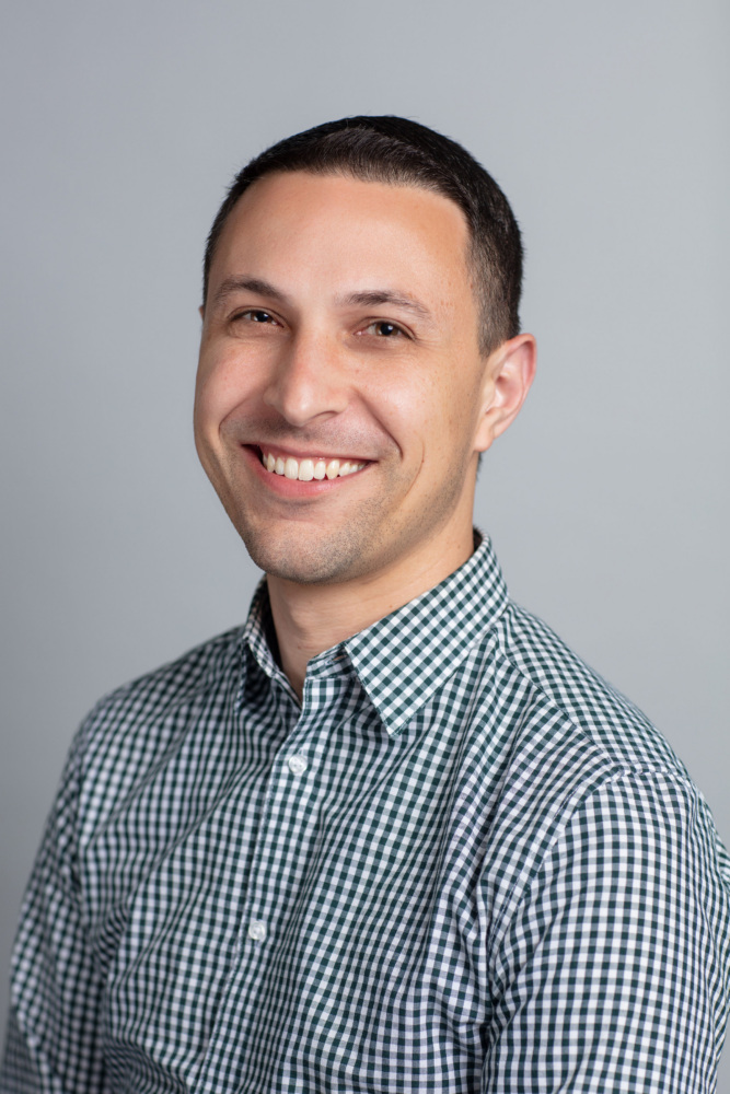 A man with short dark hair is smiling at the camera, wearing a green and white checkered shirt, against a plain gray background.