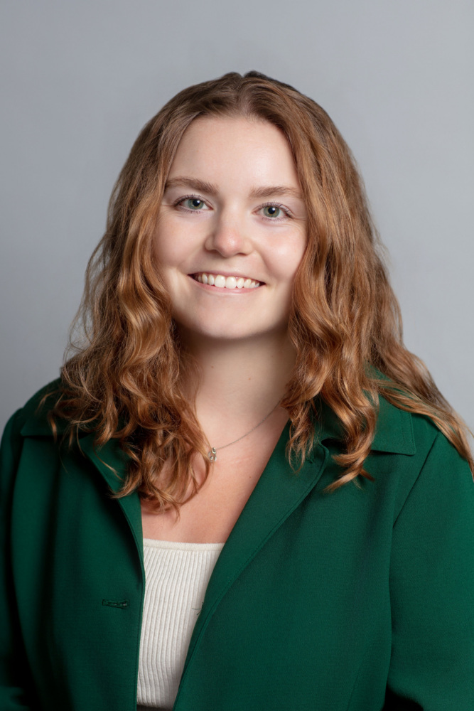 A woman with wavy light brown hair, wearing a green blazer over a beige top, smiles at the camera against a plain light gray background.