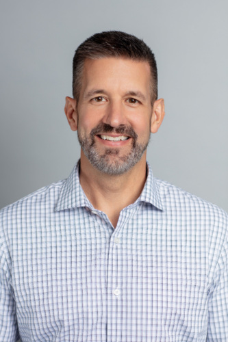 A man with short dark hair and a trimmed beard is smiling, wearing a light-colored, checkered button-up shirt against a plain gray background.