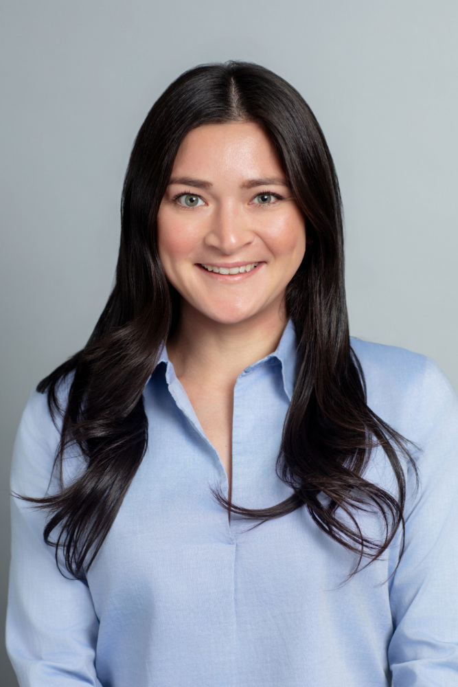A woman with long dark hair wearing a light blue button-up shirt smiles at the camera against a plain grey background.