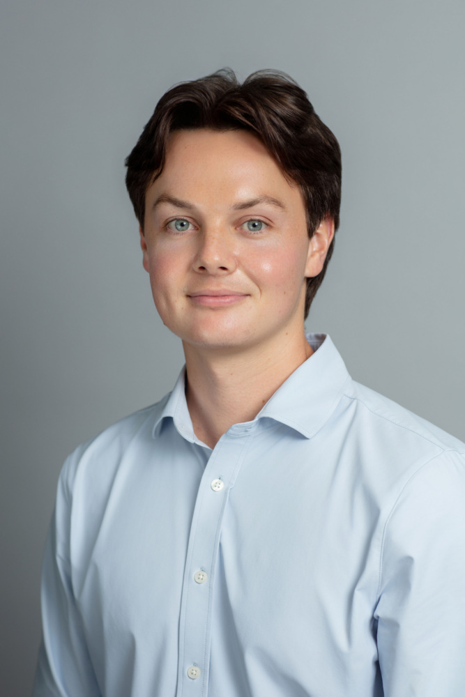 A young man with short brown hair wearing a light blue button-up shirt poses in front of a plain grey background.