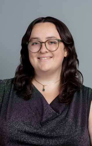 Person with long dark hair and glasses, wearing a sleeveless textured top, a necklace with a green pendant, and smiling in front of a plain gray background.