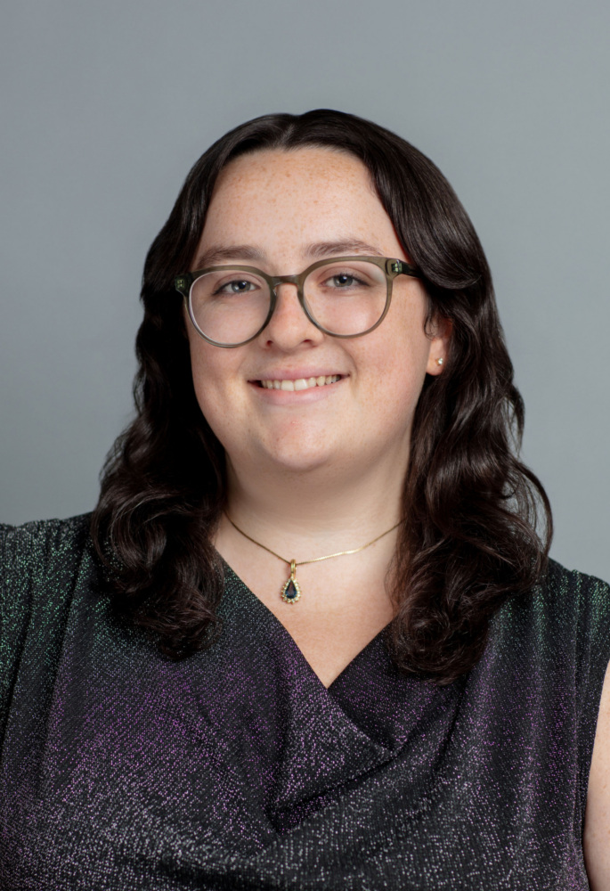 Person with long dark hair and glasses, wearing a sleeveless textured top, a necklace with a green pendant, and smiling in front of a plain gray background.