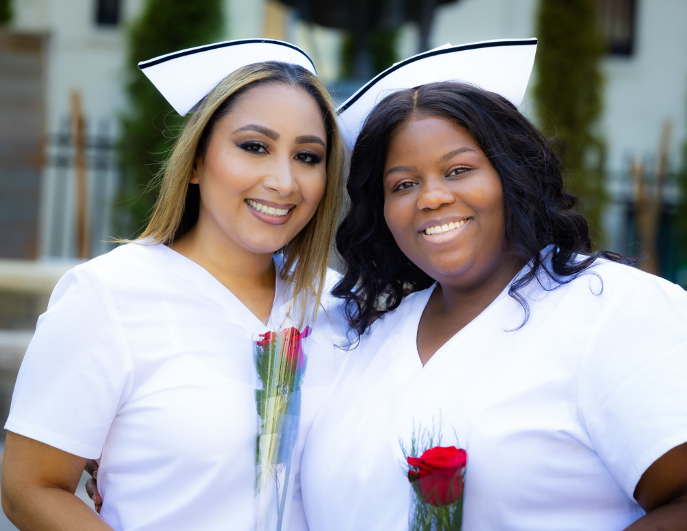 Two women wearing white uniforms and nurse caps smile and hold red rose flowers, standing outdoors in front of a blurred background.