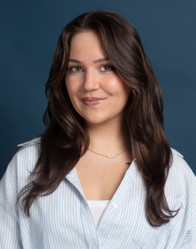 A woman with long brown hair, wearing a blue and white striped shirt and a gold necklace, smiles in front of a plain blue background.