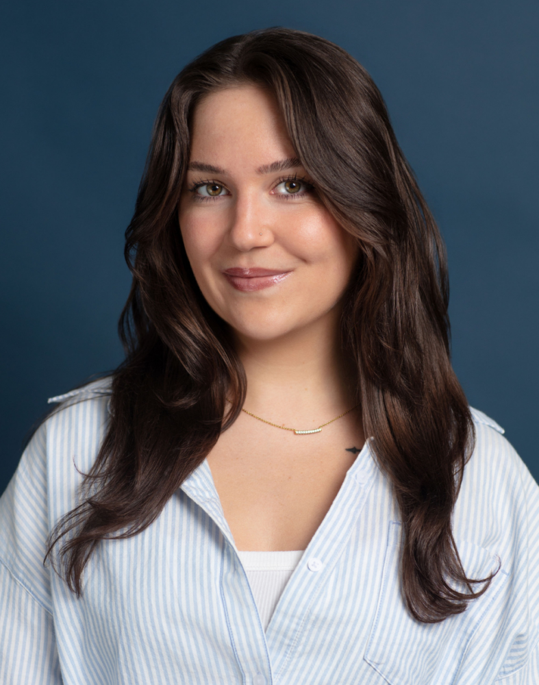 A woman with long brown hair, wearing a blue and white striped shirt and a gold necklace, smiles in front of a plain blue background.