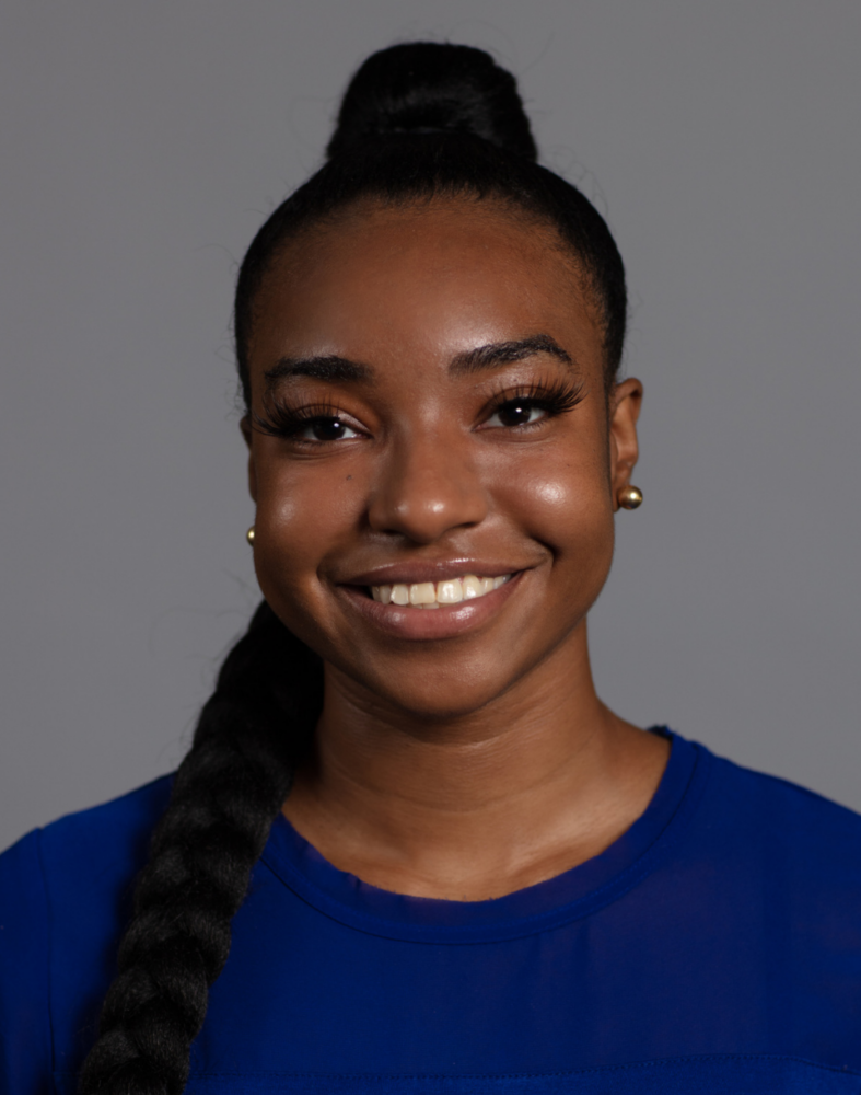 A woman with a braided ponytail, wearing a blue top and gold earrings, smiles against a plain gray background.