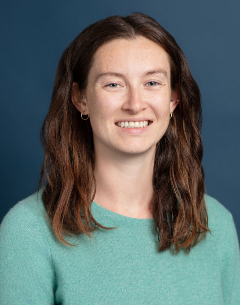 A woman with long brown hair and fair skin, wearing a mint green sweater, smiles at the camera against a plain blue background.