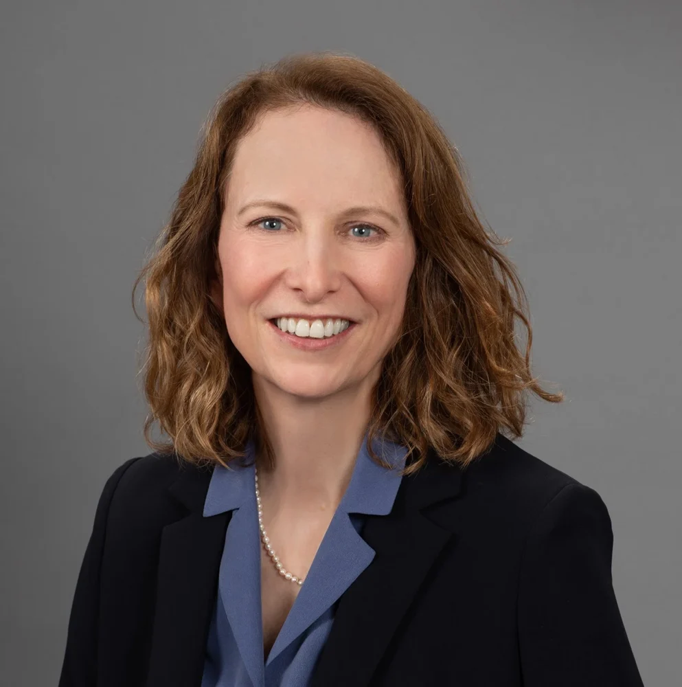 A woman with shoulder-length wavy brown hair, wearing a navy blazer, blue blouse, and a pearl necklace, smiles in front of a plain gray background.