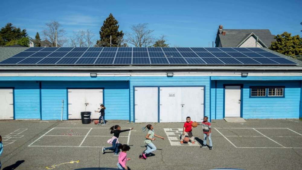Children play basketball and run in a schoolyard in front of a blue building with solar panels on the roof under a clear sky.