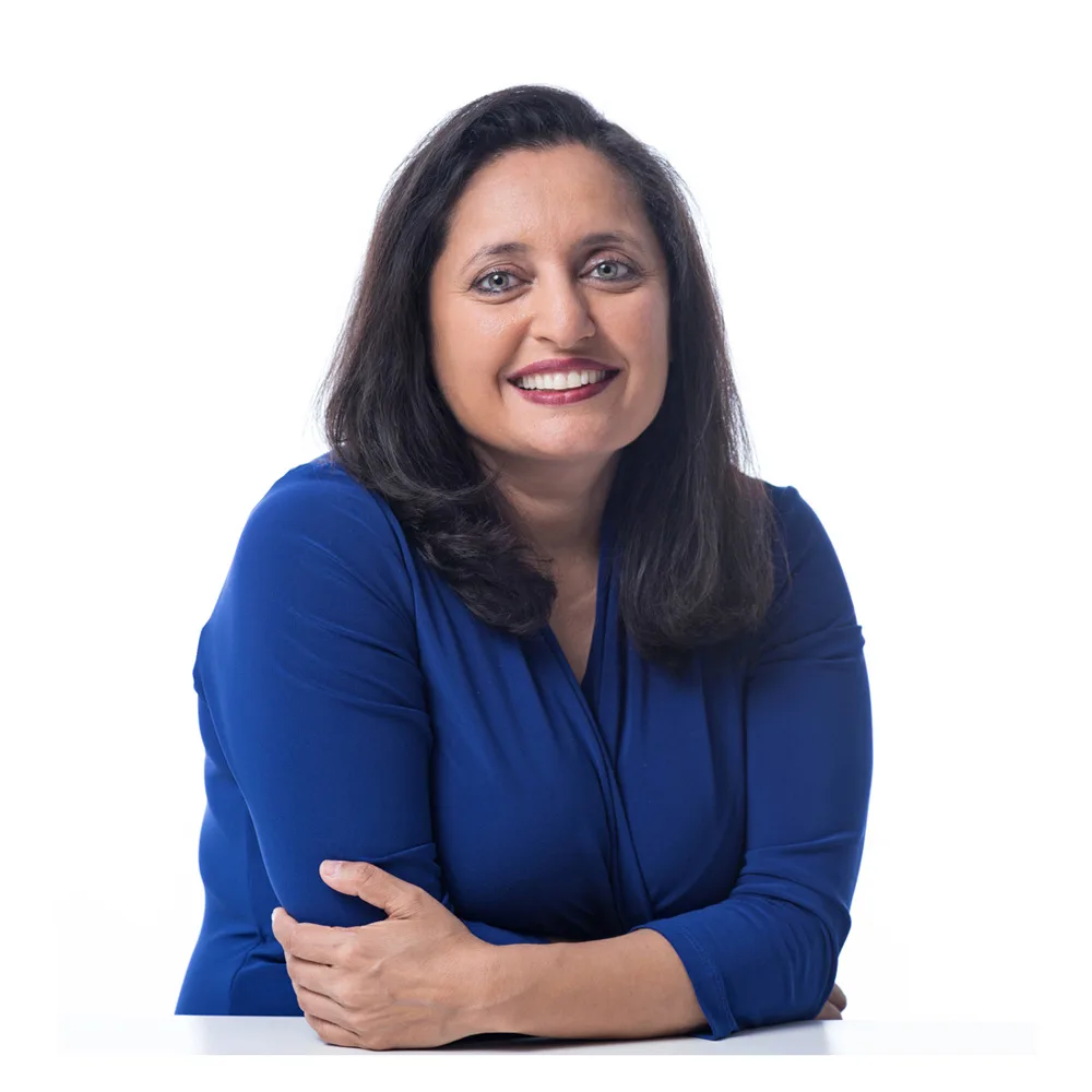 Woman with shoulder-length dark hair and a blue long-sleeve top sits at a white table, smiling at the camera against a plain white background.
