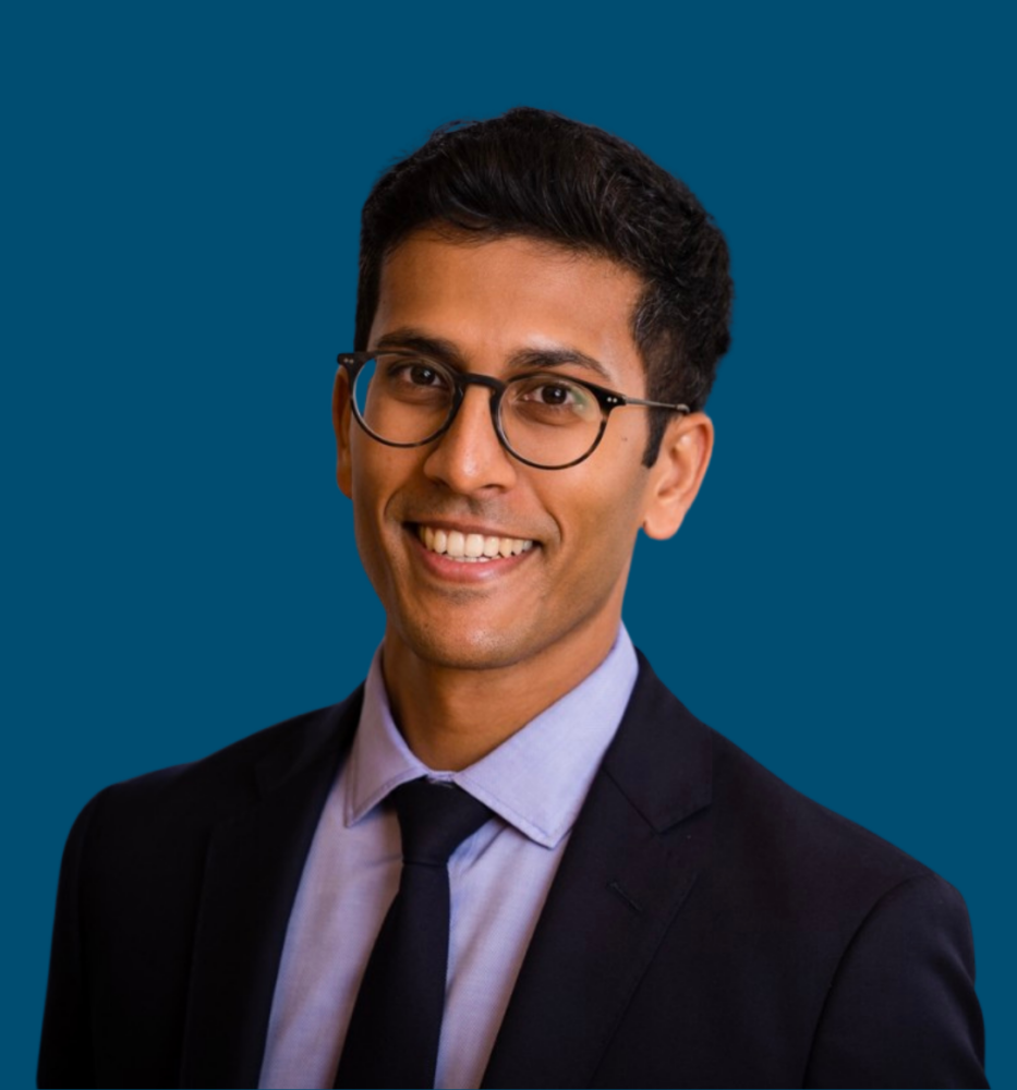 A man wearing glasses, a dark suit, and a light blue shirt with a tie, smiling in front of a solid blue background.