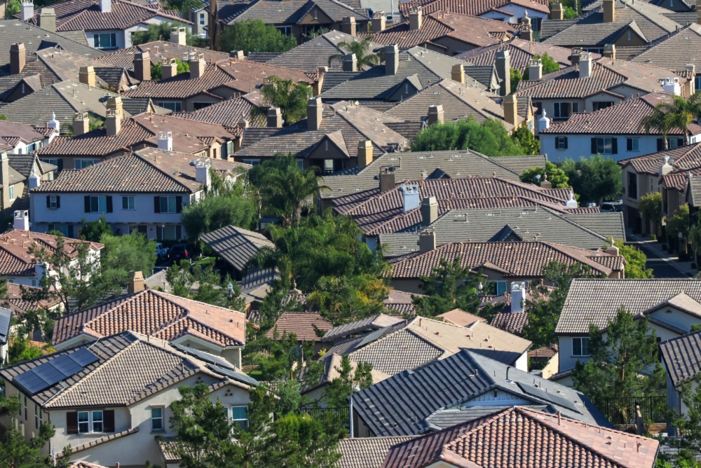 Rows of closely packed suburban houses with tiled roofs, surrounded by trees and greenery, viewed from above in daylight.