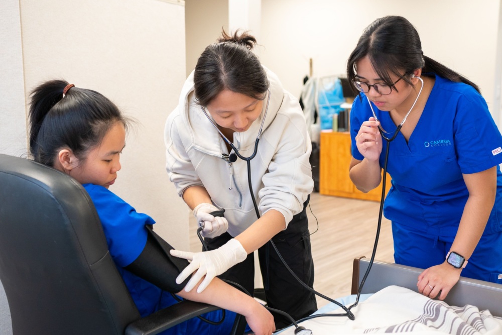 Two healthcare workers check a patient’s blood pressure in a clinical setting, both using stethoscopes and wearing blue uniforms, while the patient sits in a chair.