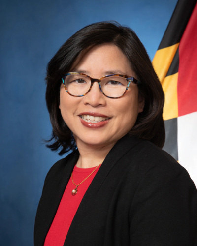 A woman with glasses and shoulder-length dark hair smiles, wearing a black blazer and red top, in front of a blue backdrop and a partial flag.
