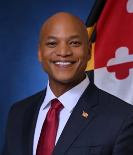 A man in a suit and red tie smiles in front of a Maryland state flag.