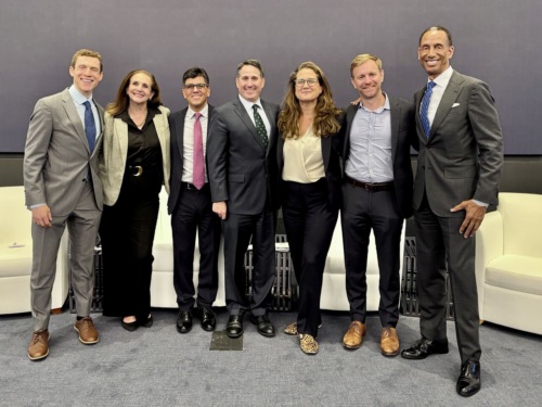 Seven people in business attire stand side by side, smiling, in front of a gray wall and white couches.