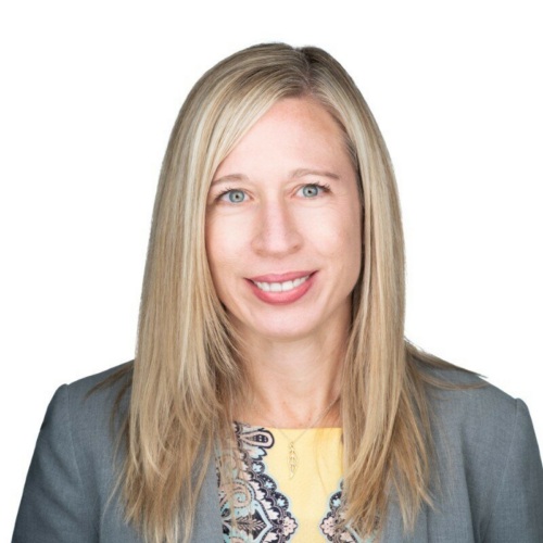 Woman with straight blonde hair wearing a grey blazer and patterned top, smiling at the camera against a plain white background.
